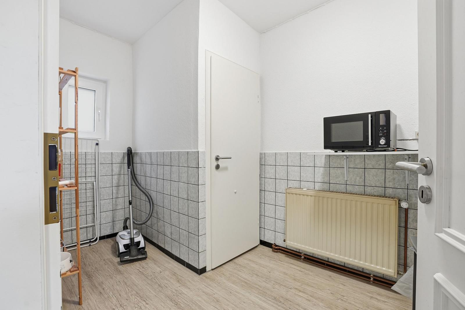 Kitchen with microwave, radiator, and vacuum cleaner. Tiled wall and wooden floor.