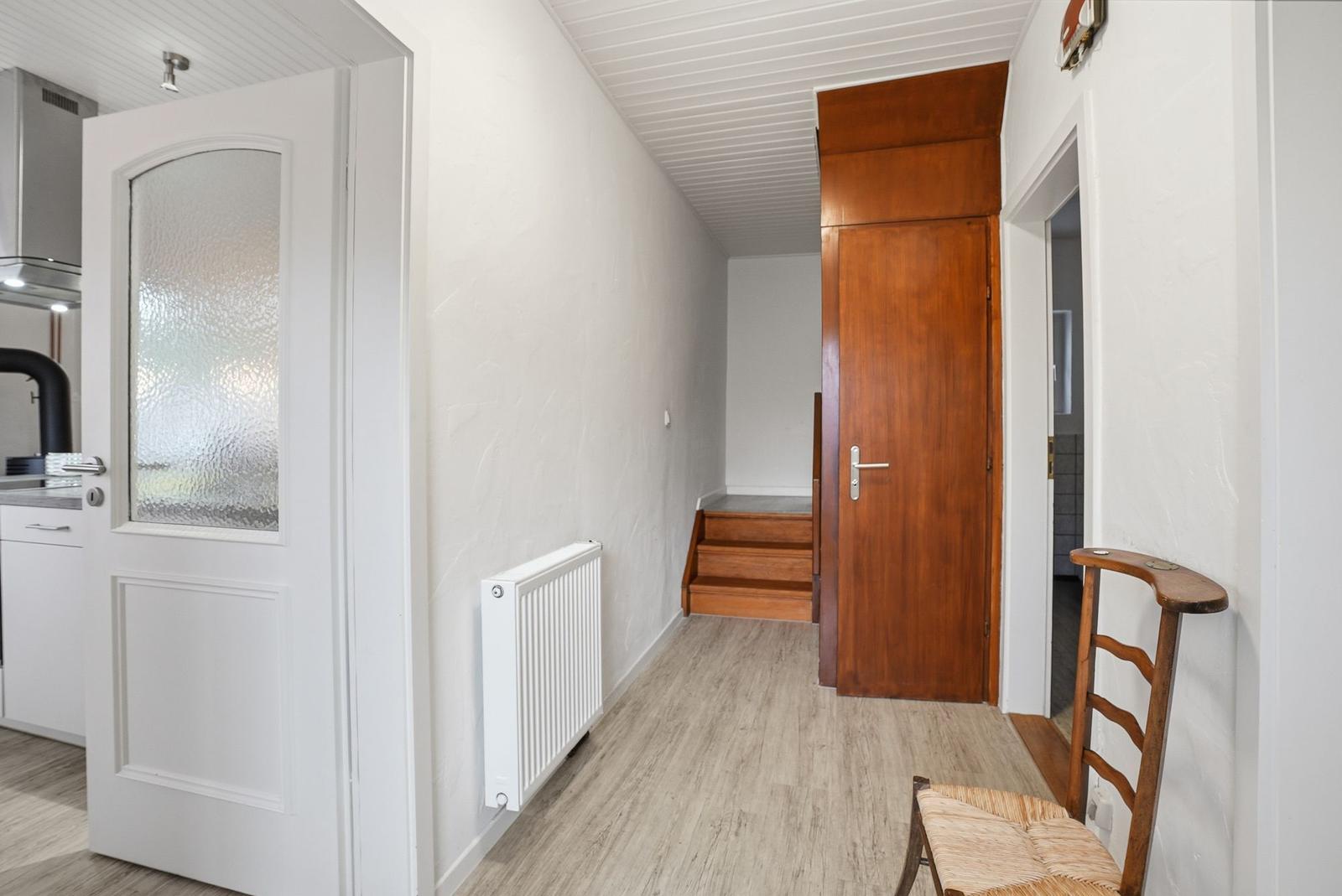 Hallway with wooden stairs, door, and chair. Kitchen visible on the left.