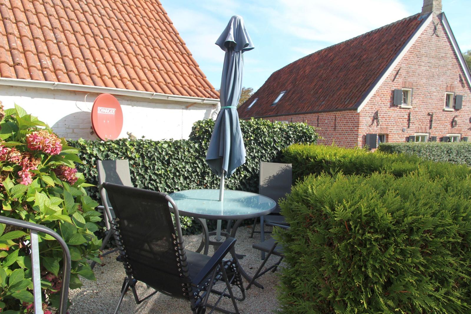 Garden with table, chairs, and closed umbrella between hedges and houses.