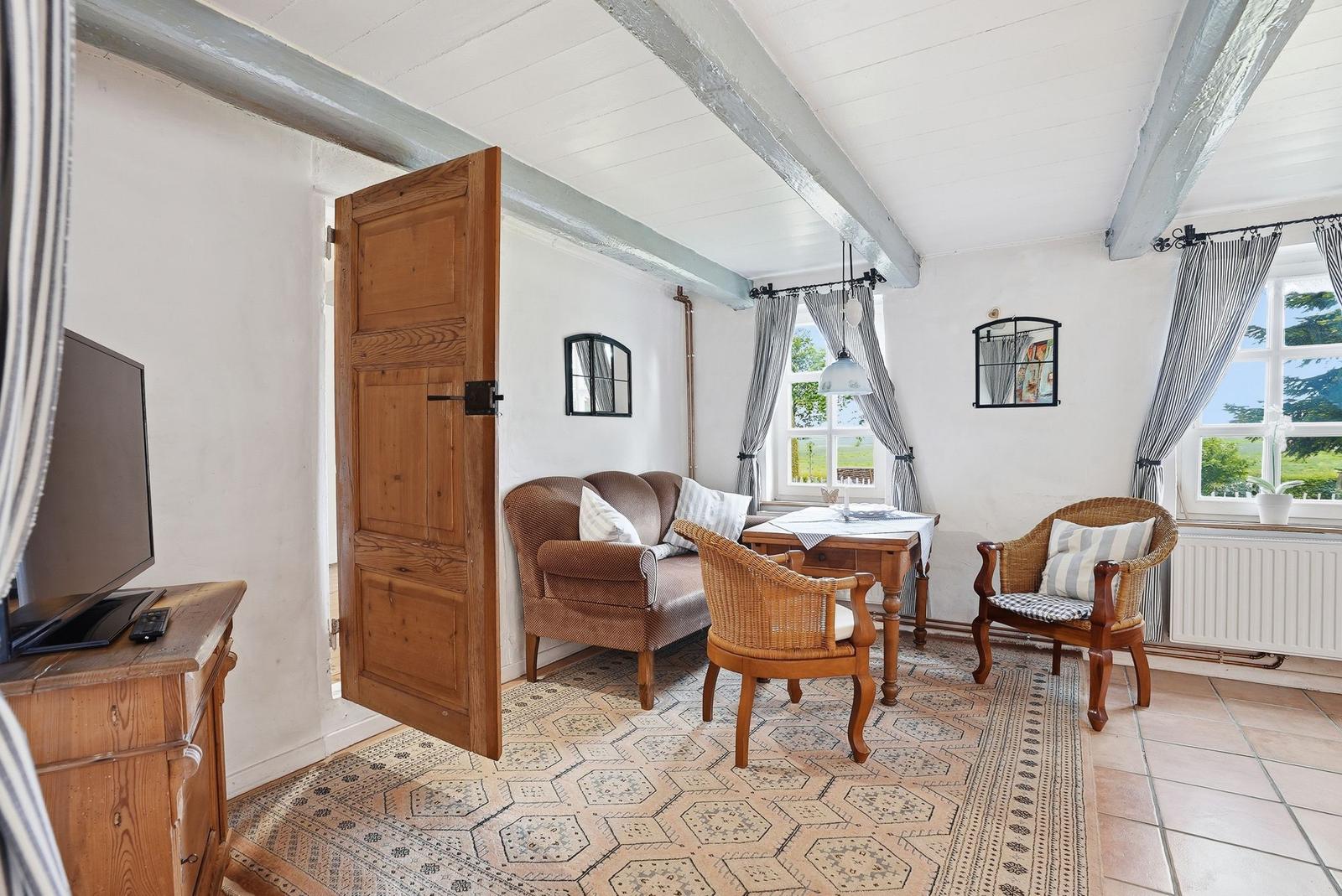 Wohnzimmer mit Holztisch, Stühlen und Sofa. Holzbalken an der Decke und Fenster mit Gardinen.