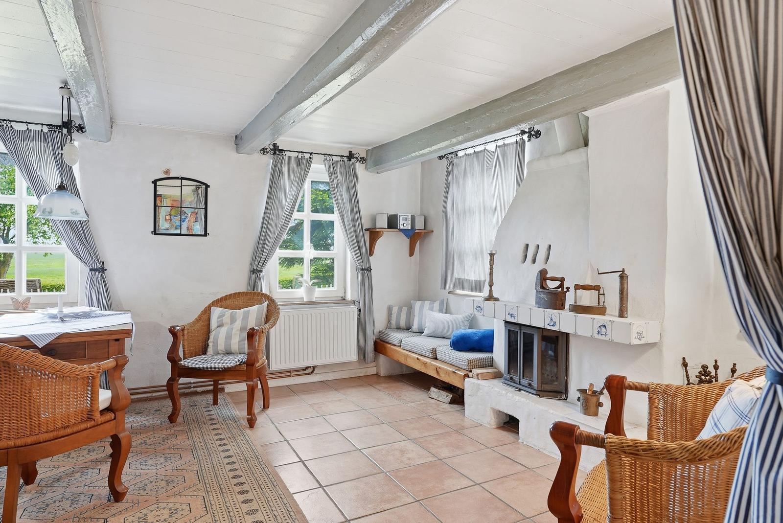 Raum mit Holzbalken, Kamin und Sitzgelegenheiten. Fenster mit Vorhängen und Fliesenboden.