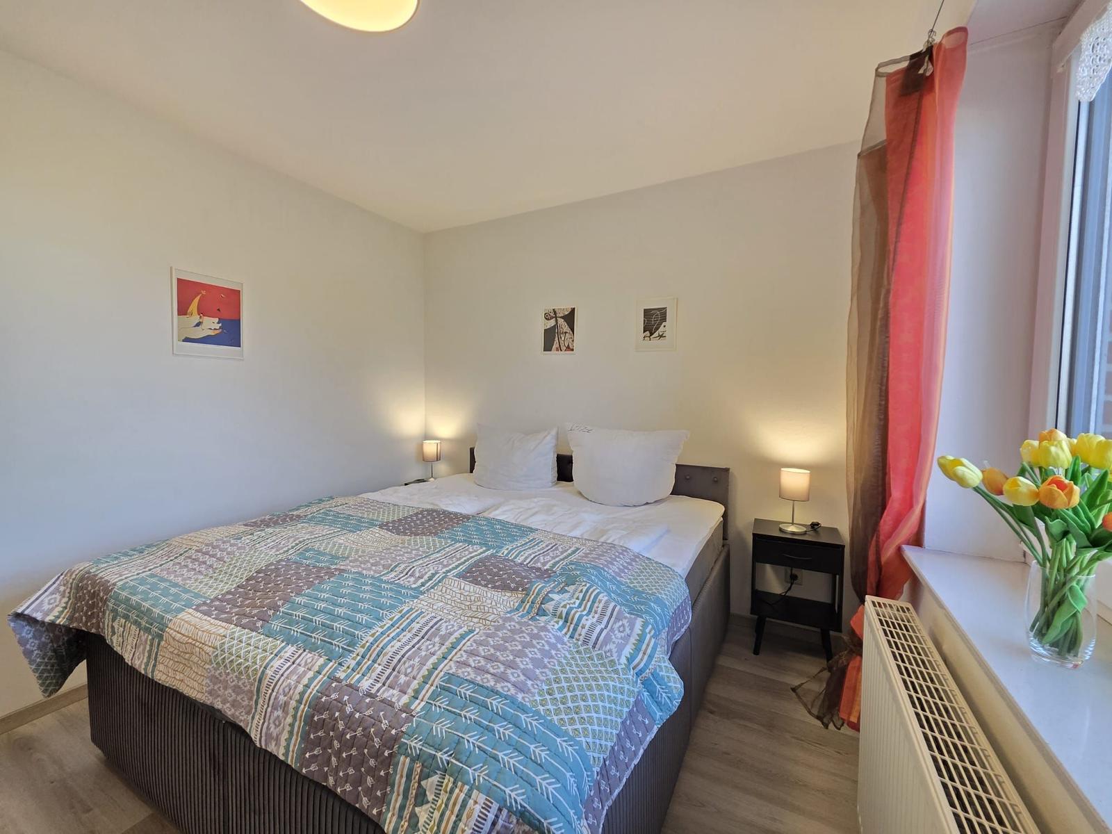 Schlafzimmer mit Doppelbett, Nachttischen und Blumen auf dem Fensterbrett.