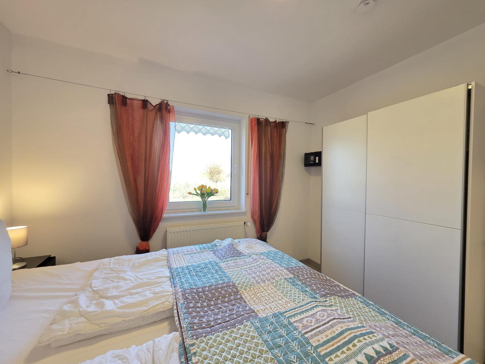 Schlafzimmer mit Bett, Fenster und Schrank. Vor dem Fenster steht eine Vase mit Blumen.