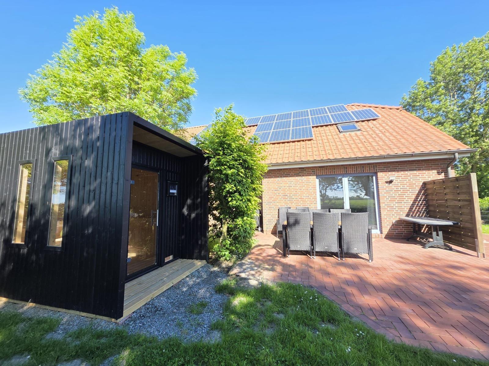 Schwarzes Holzhaus neben einem Backsteinhaus mit Solaranlage auf Dach und Terrasse mit Gartenmöbeln.