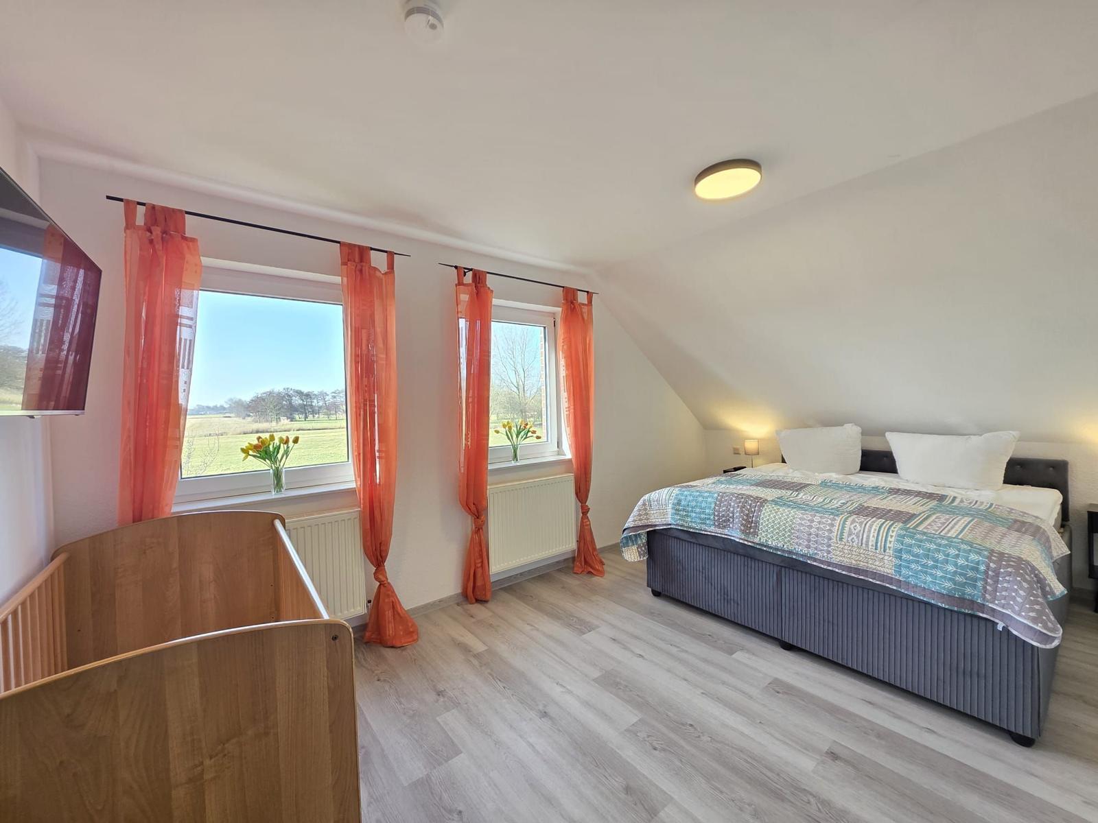 Schlafzimmer mit Bett, Babybett und Fenstern mit orangefarbenen Vorhängen.