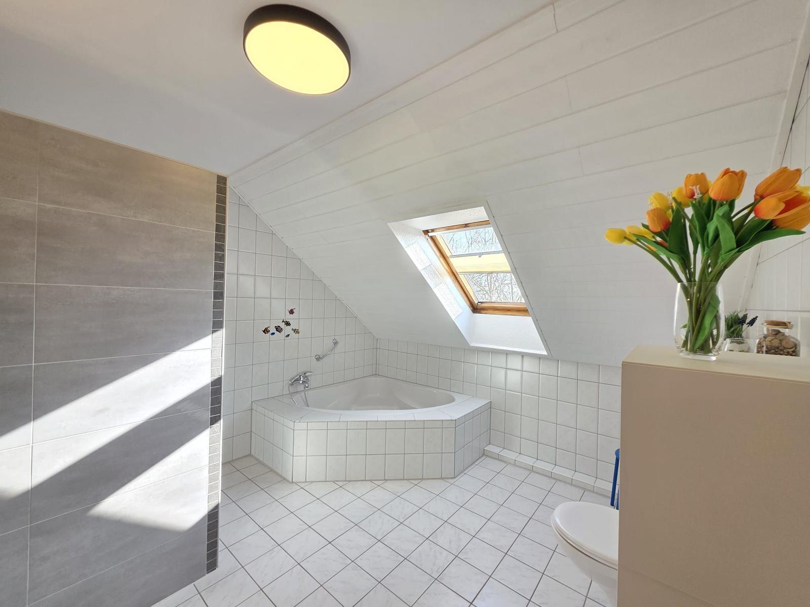 Dusche mit Duschwand, Badewanne und Dachfenster. Auf dem Waschtisch steht eine Vase mit Tulpen.
