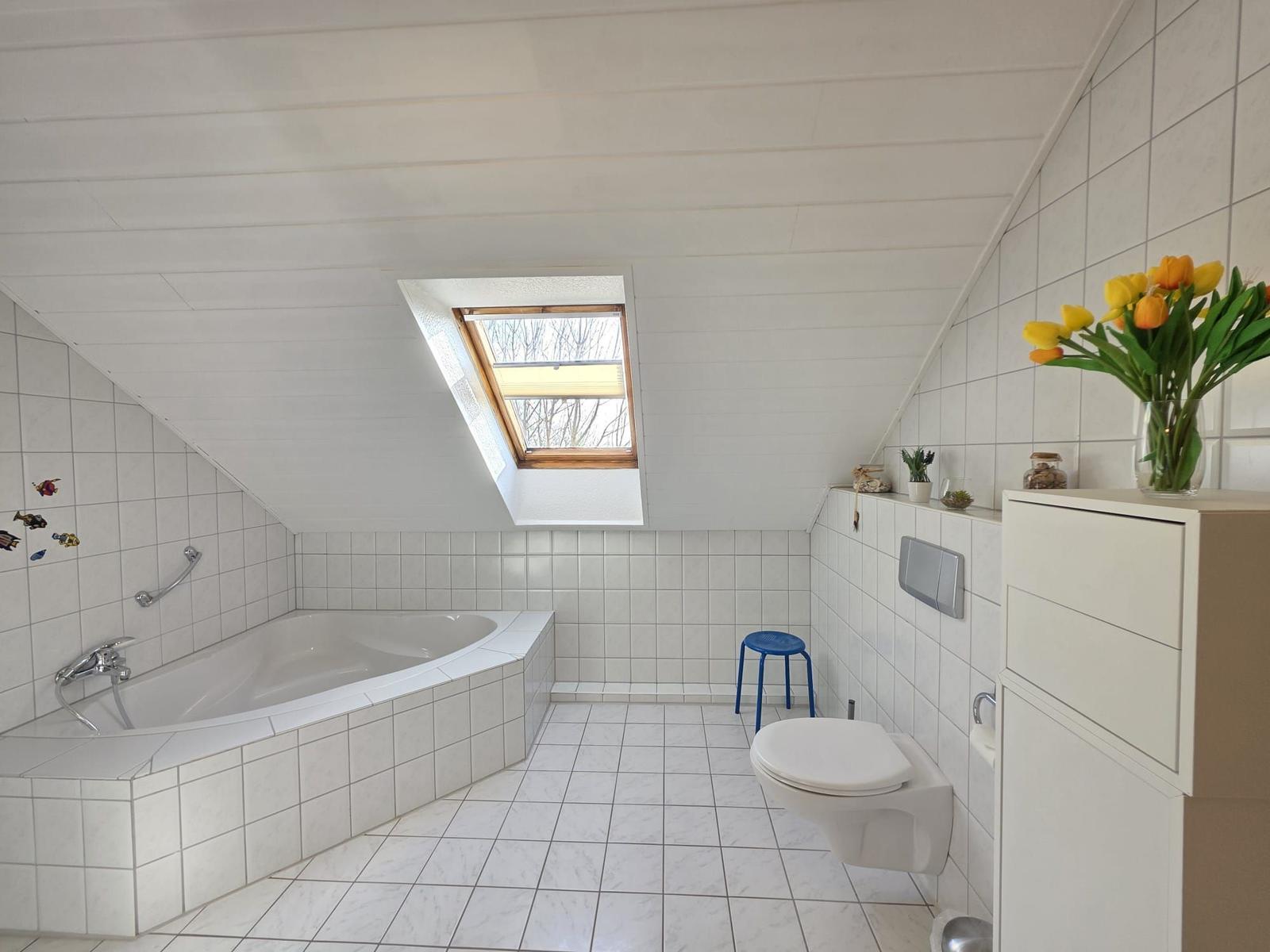 Badezimmer mit Badewanne, Toilette und Dachfenster. Weiße Fliesen und ein Blumenstrauß.