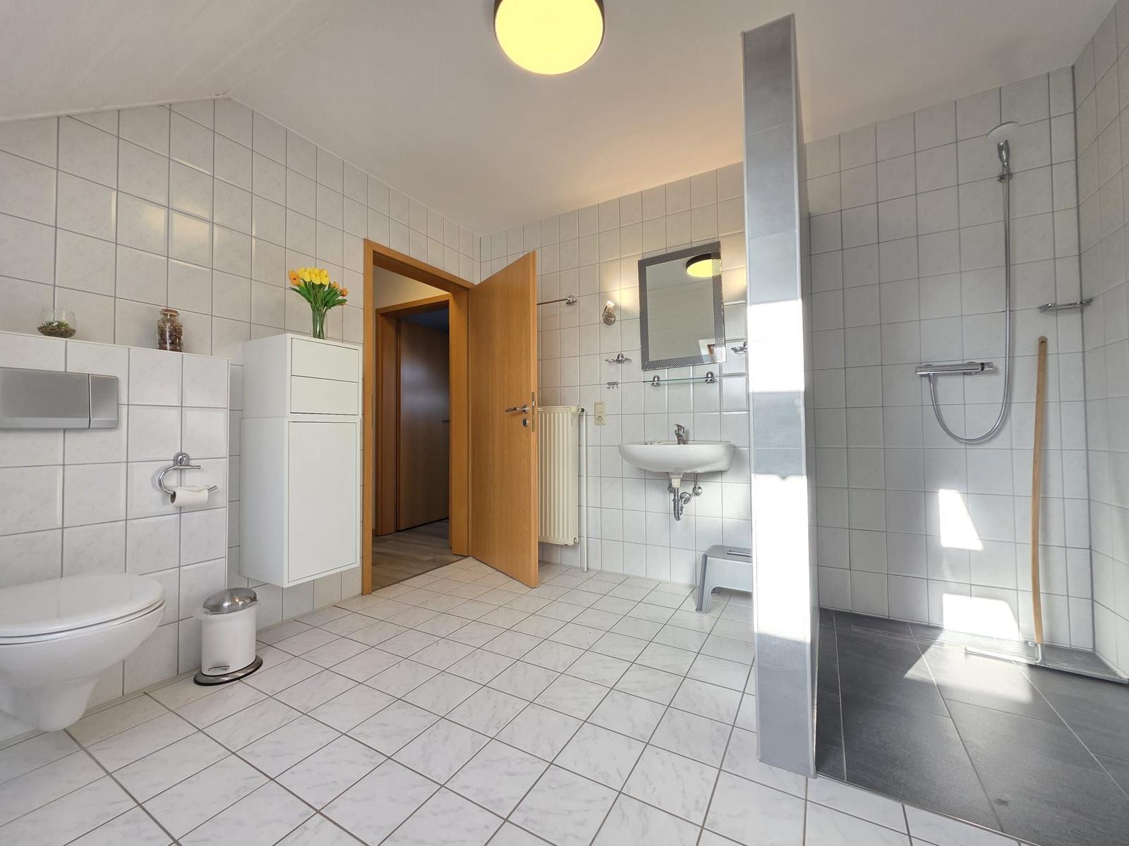 Badezimmer mit Dusche, Waschbecken und Toilette. Weiße Fliesen und Holztür.