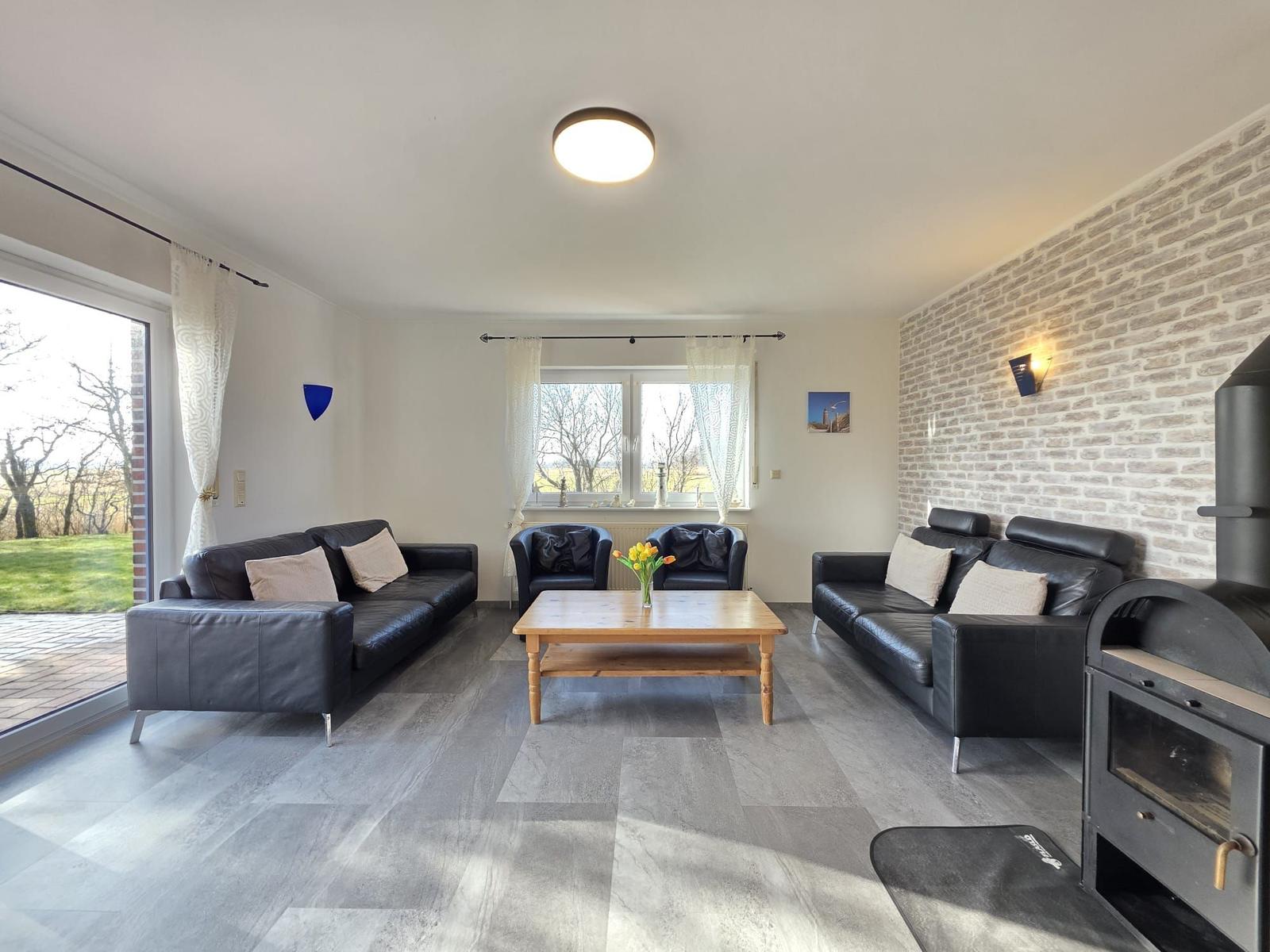Wohnzimmer mit zwei schwarzen Sofas, Holztisch und Kaminofen. Durchgang zur Terrasse.