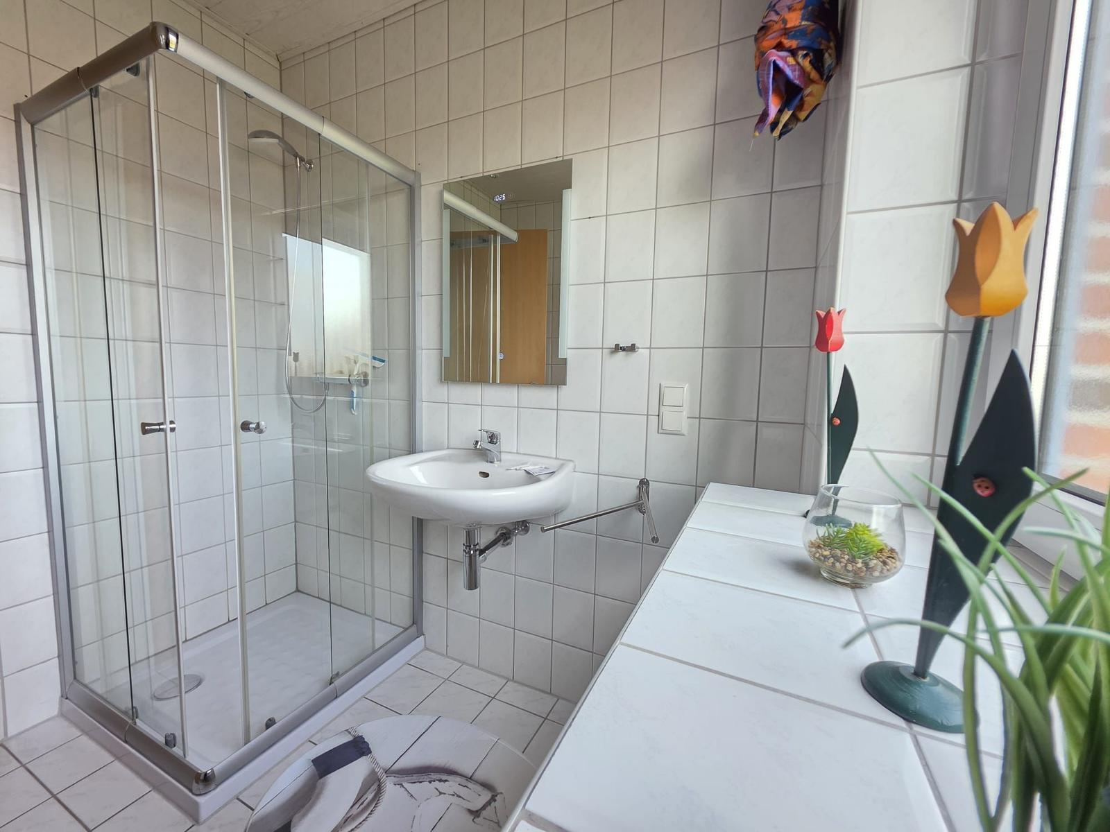 Badezimmer mit Dusche, Waschbecken und Fenster mit Pflanzen.