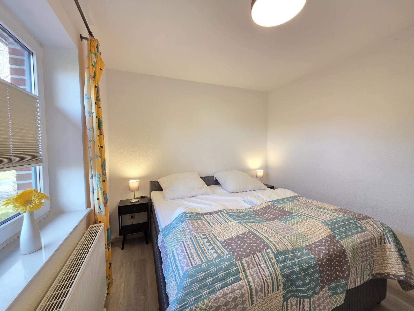 Schlafzimmer mit Bett, Nachttisch und Fenster mit Vorhang.
