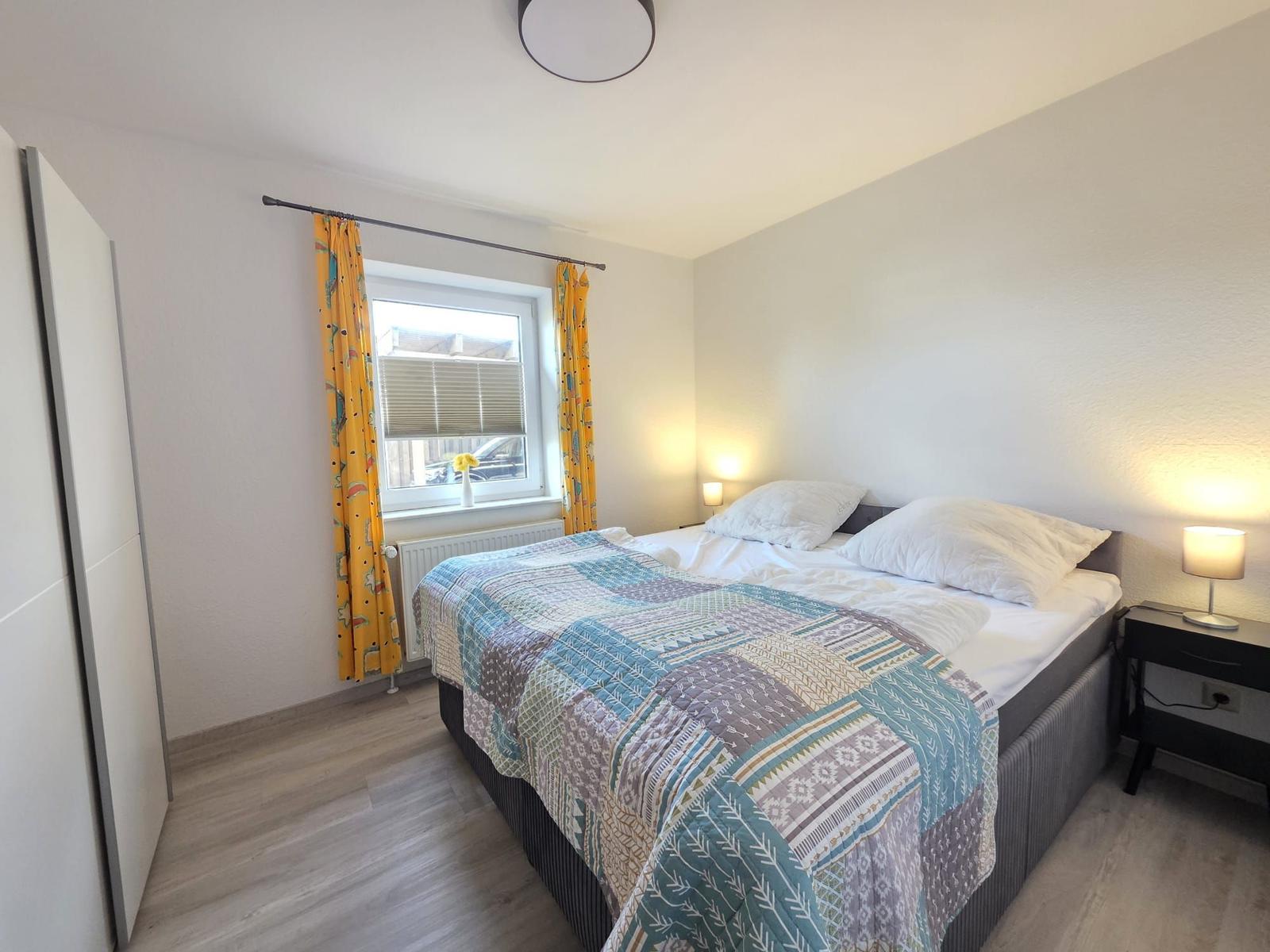 Schlafzimmer mit Bett, Nachttisch und Fenster mit gelben Vorhängen.