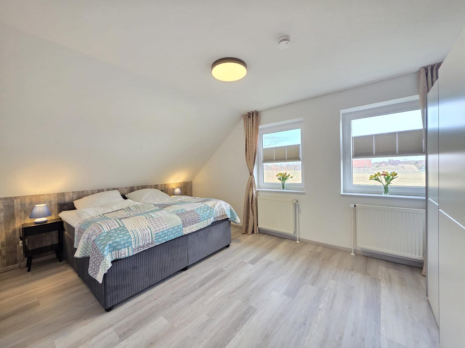 Schlafzimmer mit Doppelbett, zwei Fenstern und Holzboden.