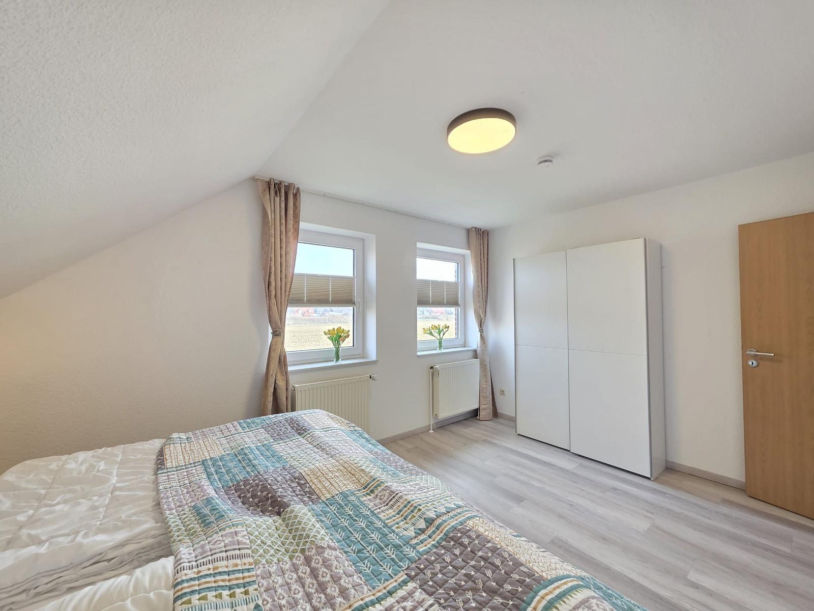 Schlafzimmer mit Dachschräge, zwei Fenstern, weißem Schrank und Holztür.