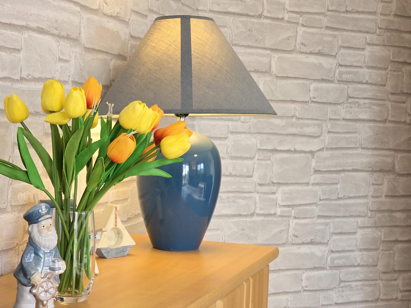 Blauer Tischleuchte mit grauem Schirm steht neben Tulpen auf Holztisch vor weißem Ziegelwand.
