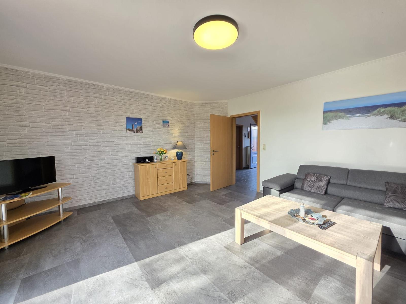 Wohnzimmer mit Sofa, Couchtisch, Fernseher und Holzmöbeln. Helle Wände und Bodenfliesen.