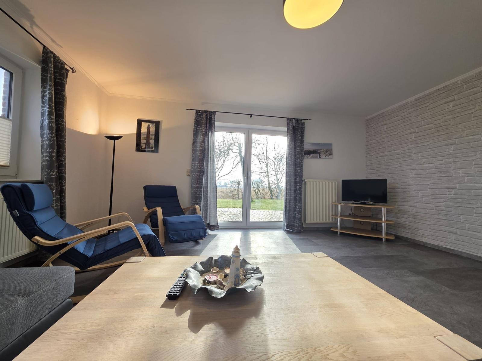 Wohnzimmer mit blauen Sesseln, TV und Terrassentür. Holztisch mit Dekoration.