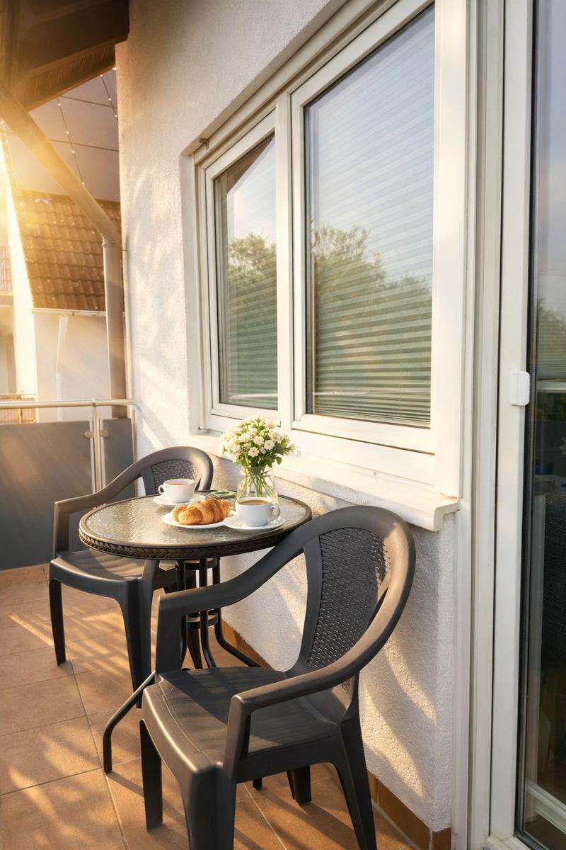 Terrasse mit Tisch, zwei Stühlen, Kaffeetasse und Croissants. Fenster mit Jalousien und Sonnenlicht.
