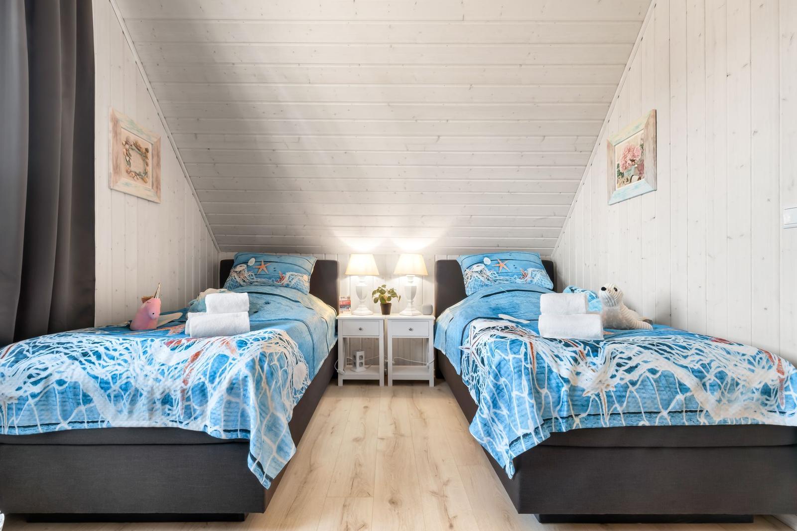 Zwei Betten mit blauer Bettwäsche, Nachttisch mit Lampe und Wandbildern in einem Dachzimmer.