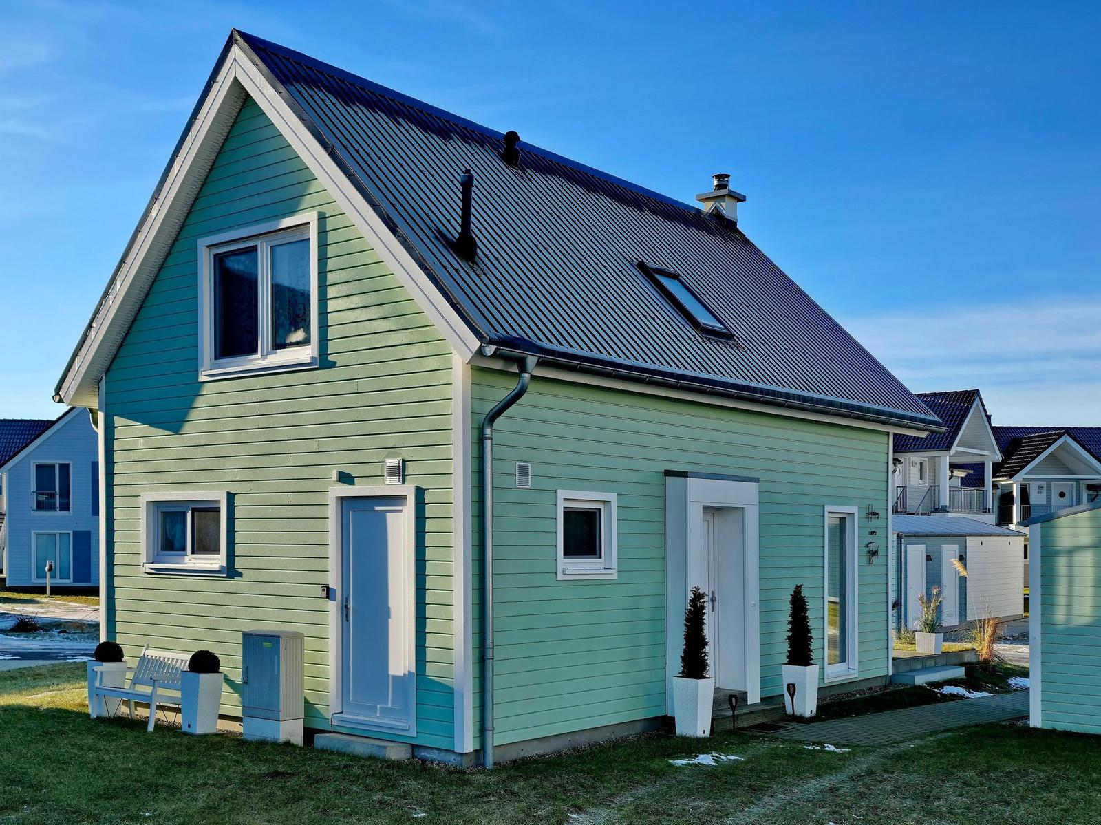 Grünes Haus mit Dachfenster und weißen Fensterrahmen. Vor dem Haus befindet sich eine kleine Terrasse.