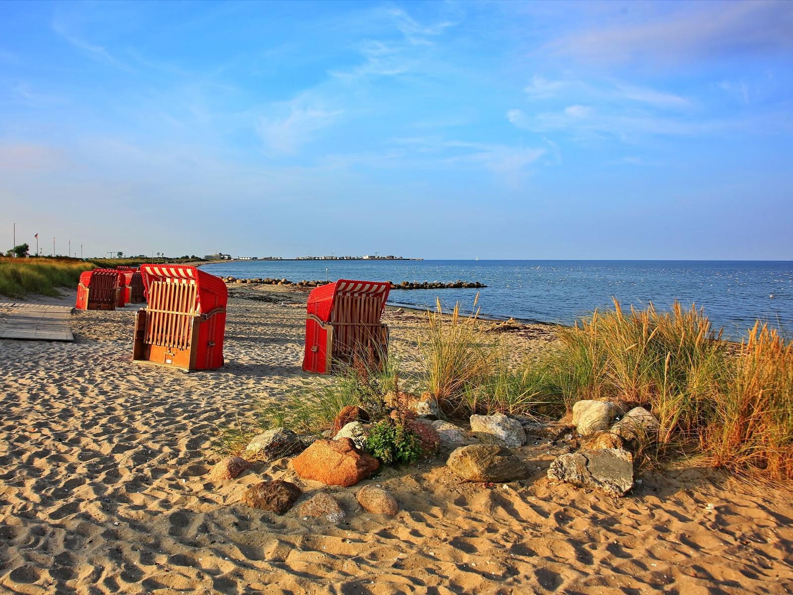 Rote Strandkörbe stehen auf sandigem Ufer, das Meer und Himmel sind klar.