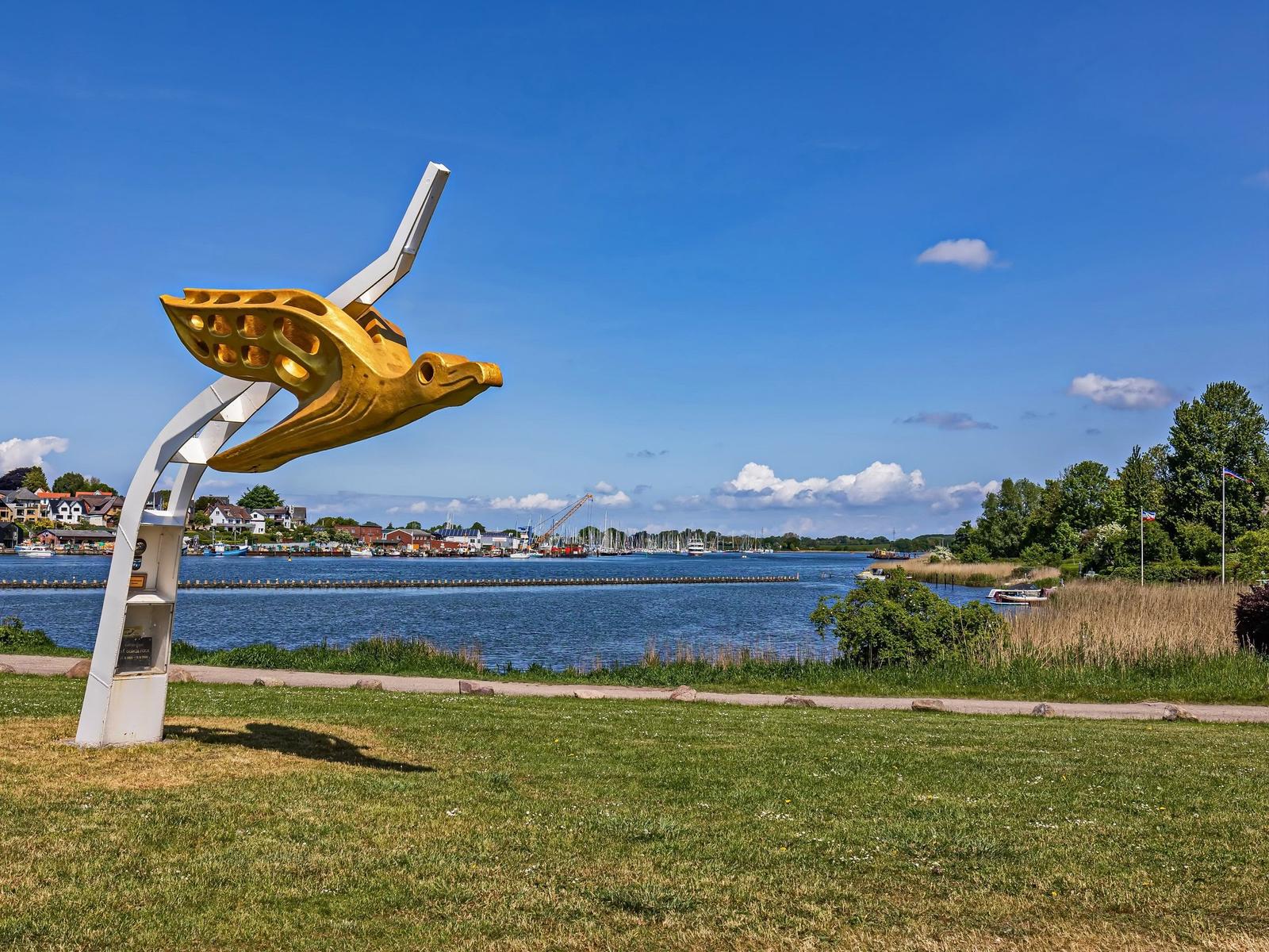 Goldene Vogelskulptur am Wasser mit Booten und Häusern im Hintergrund.