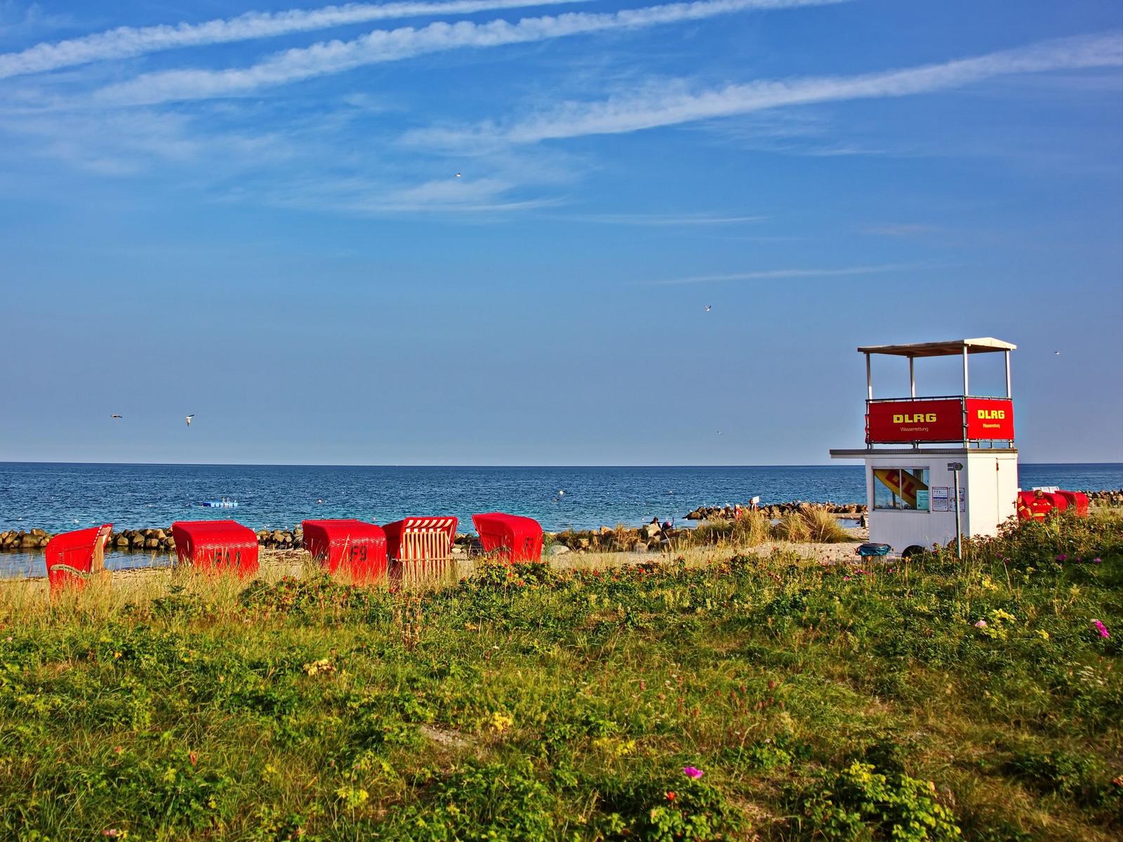 Rote Strandkörbe und ein Wachturm am Strand mit Meer und blauem Himmel.