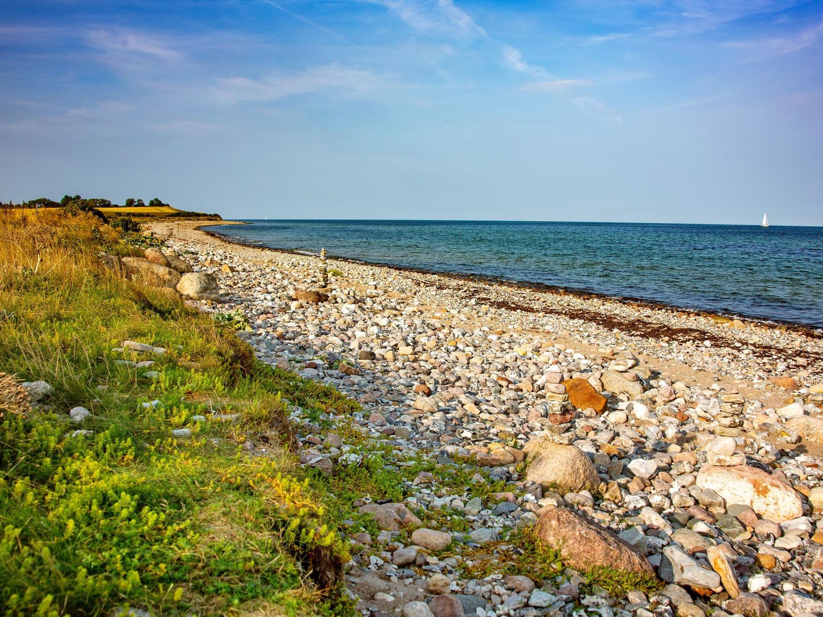 Steiniger Strand mit Gras und einem Segelboot im Hintergrund.