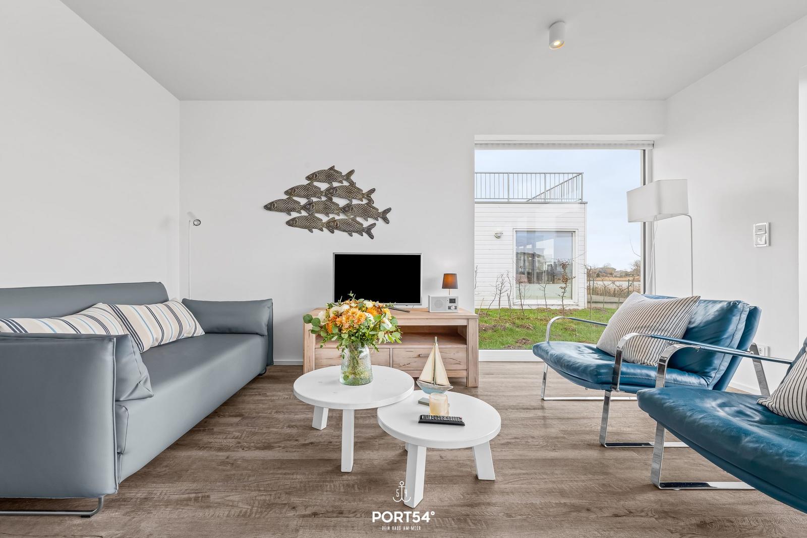 Wohnzimmer mit Sofa, Sesseln und TV. Fensterblick auf Balkon und Grünfläche.