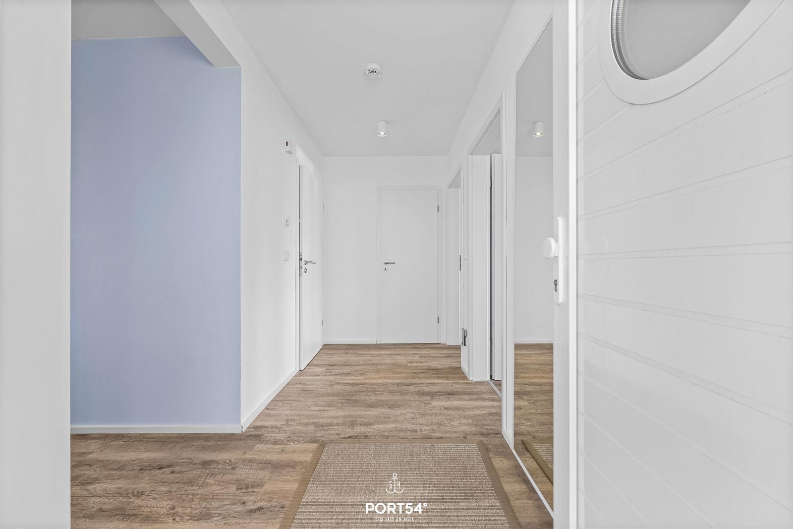 Flur mit Holzboden und weißen Wänden. Eine Tür führt in einen Raum mit runder Fensteröffnung.