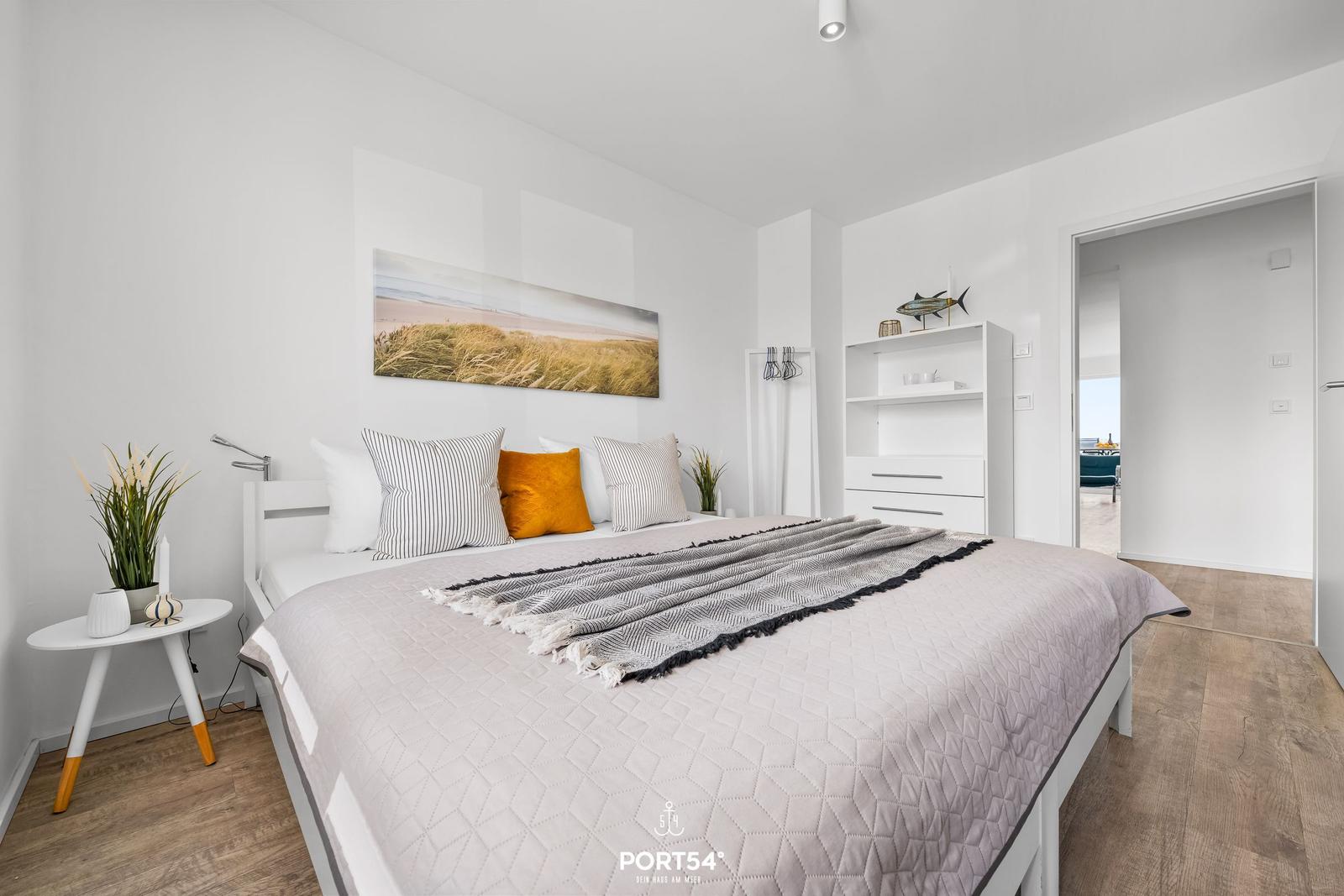 Schlafzimmer mit Bett, Nachttisch und Wandbild. Tür führt in Wohnbereich.