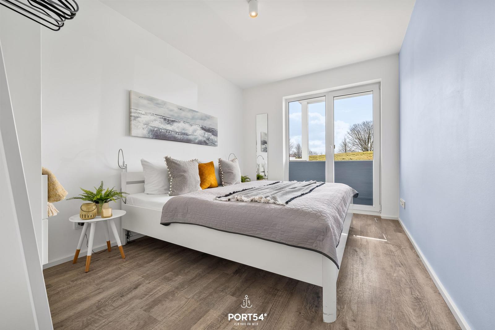 Schlafzimmer mit weißem Bett, Fenster zur Terrasse und Wandbild mit Wellenmotiv.