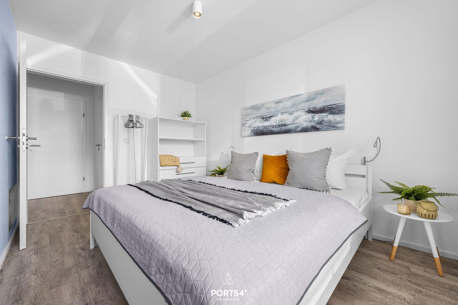 Schlafzimmer mit Bett, Nachttisch und Wandbild. Hintergrund: Tür und Kleiderständer.