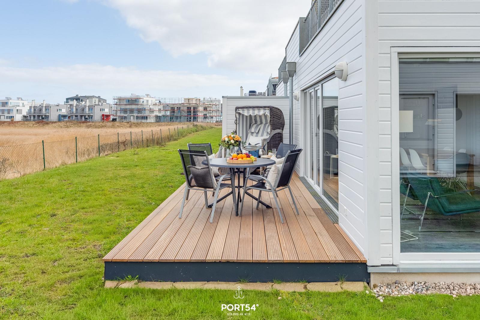 Terrasse mit Tisch, Stühlen und Liegestuhl. Grünflächen und moderne Gebäude im Hintergrund.