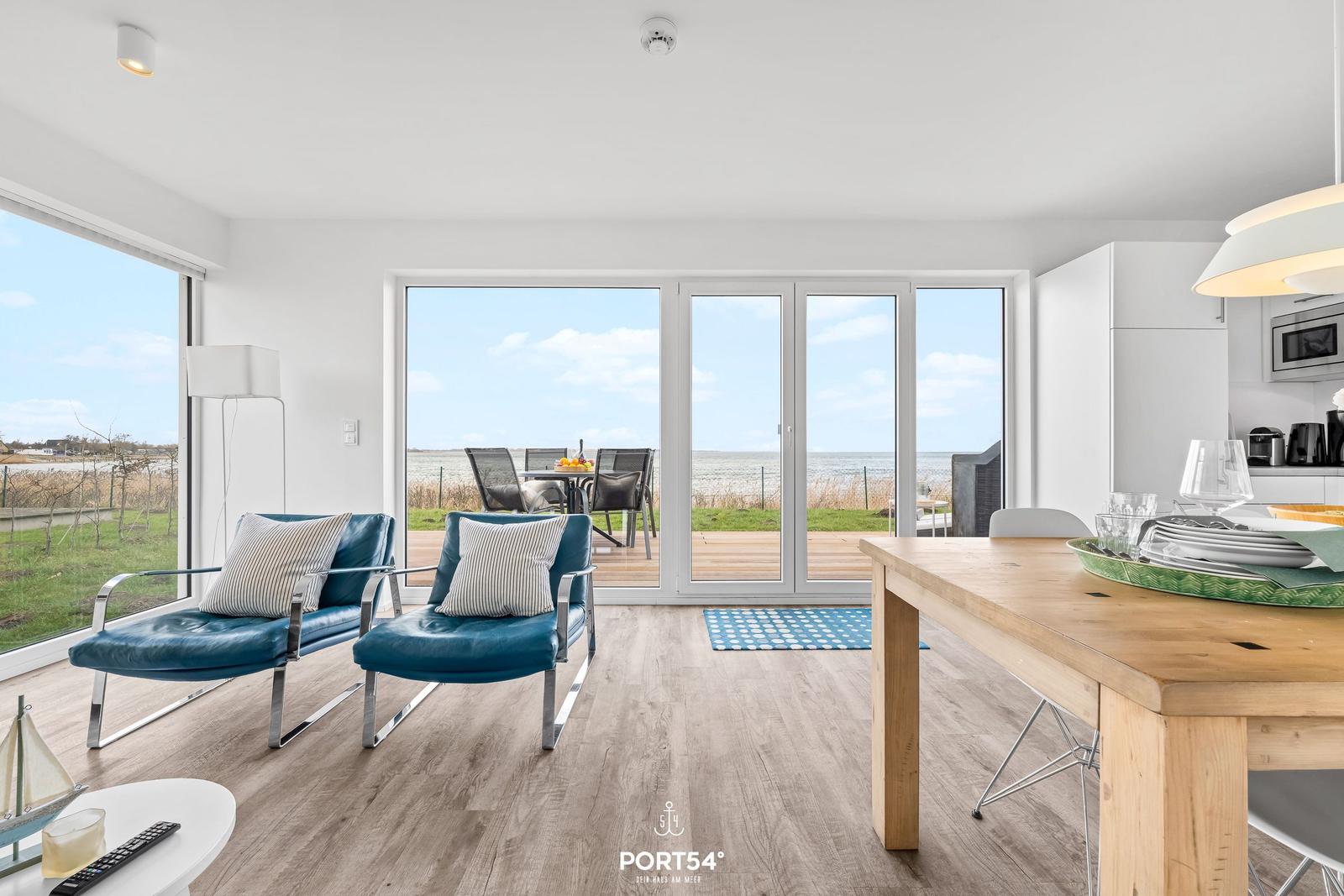 Wohn- und Essbereich mit Blick aufs Meer und Terrasse. Blaue Sessel und Holztisch.