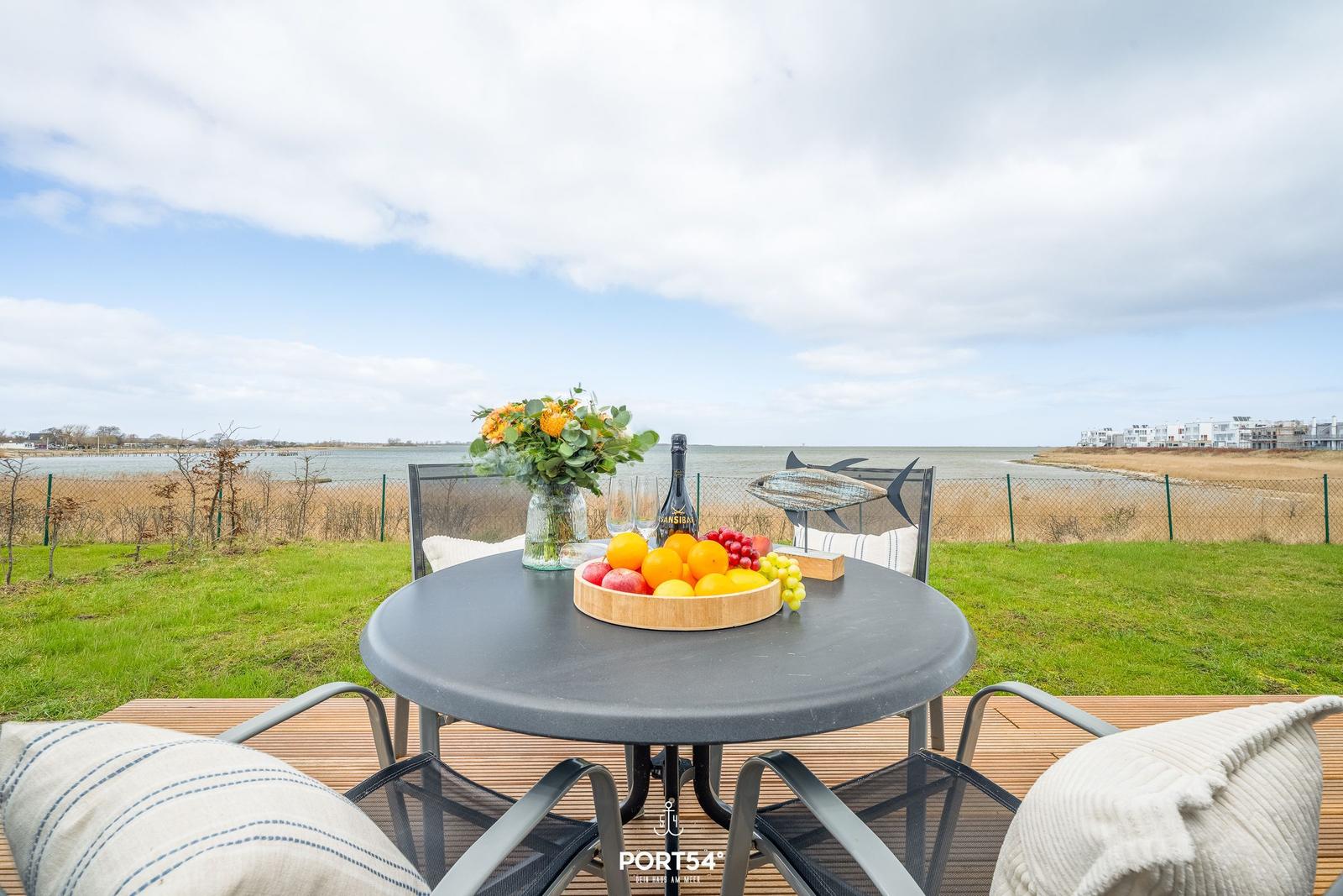Runder Tisch mit Früchten und Blumen auf Terrasse mit Blick auf Wasser und Gras.