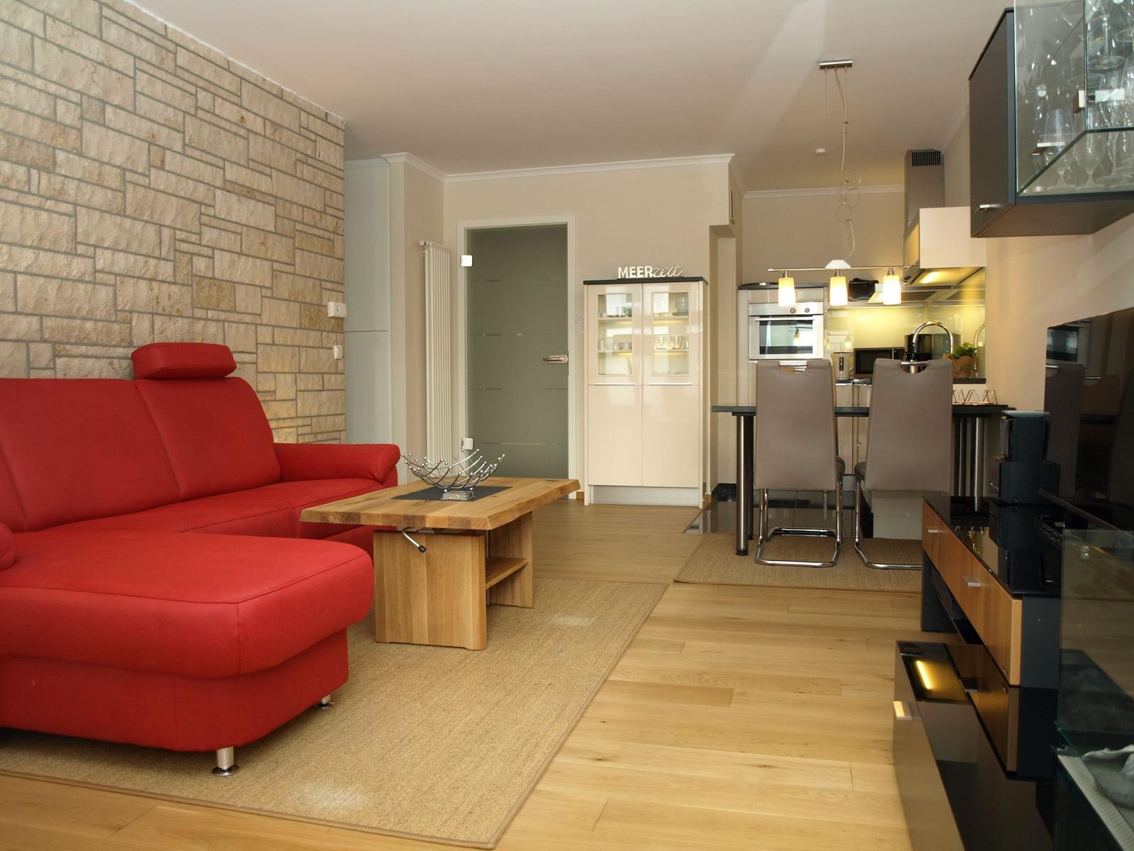 Ein Wohnzimmer mit rotem Sofa, Holztisch und Steinwand. Küche mit Essbereich im Hintergrund.