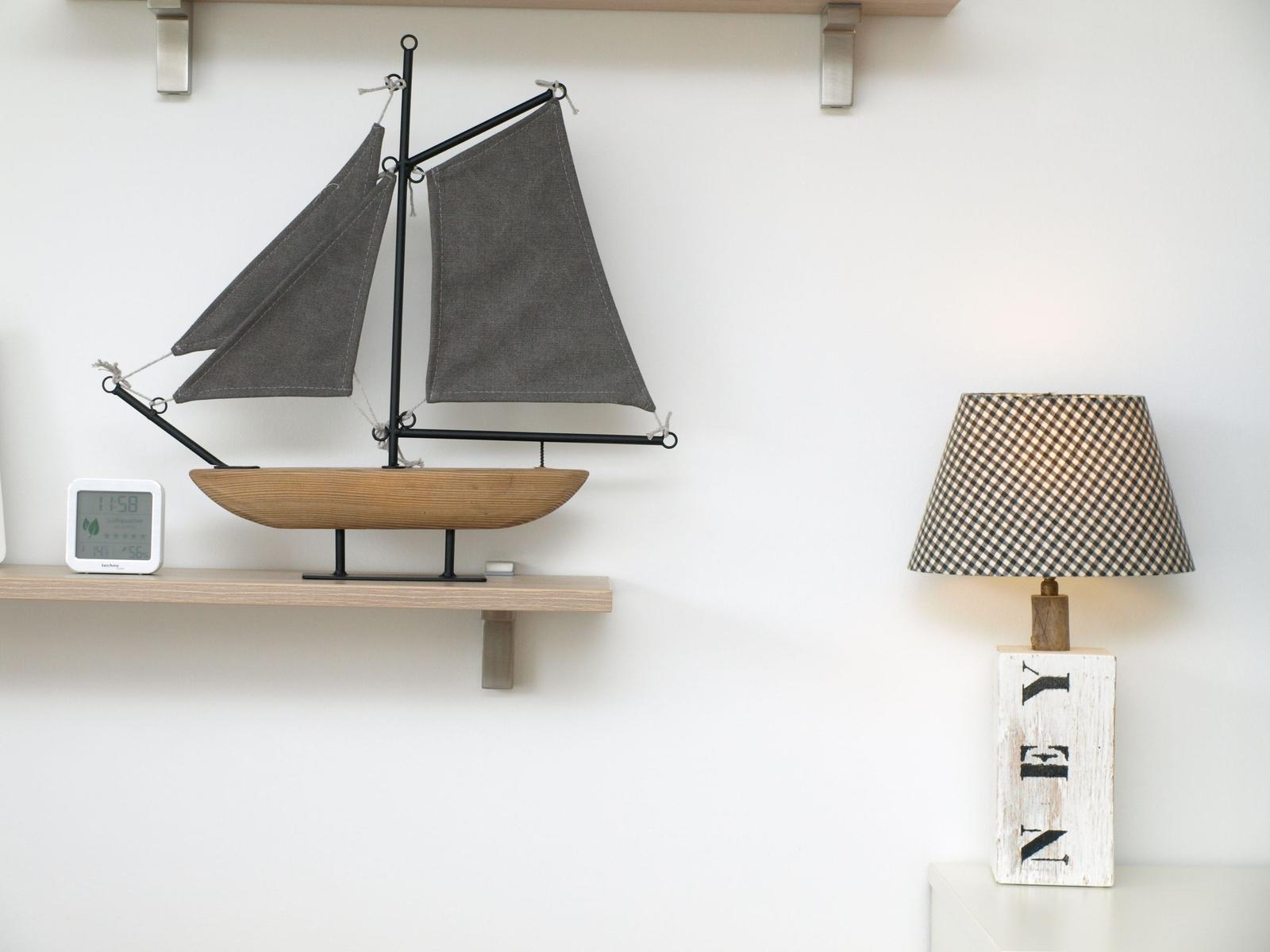 Ein Modellboot mit grauen Segeln steht auf einer Holzregal. Daneben steht eine Lampe mit kariertem Schirm.