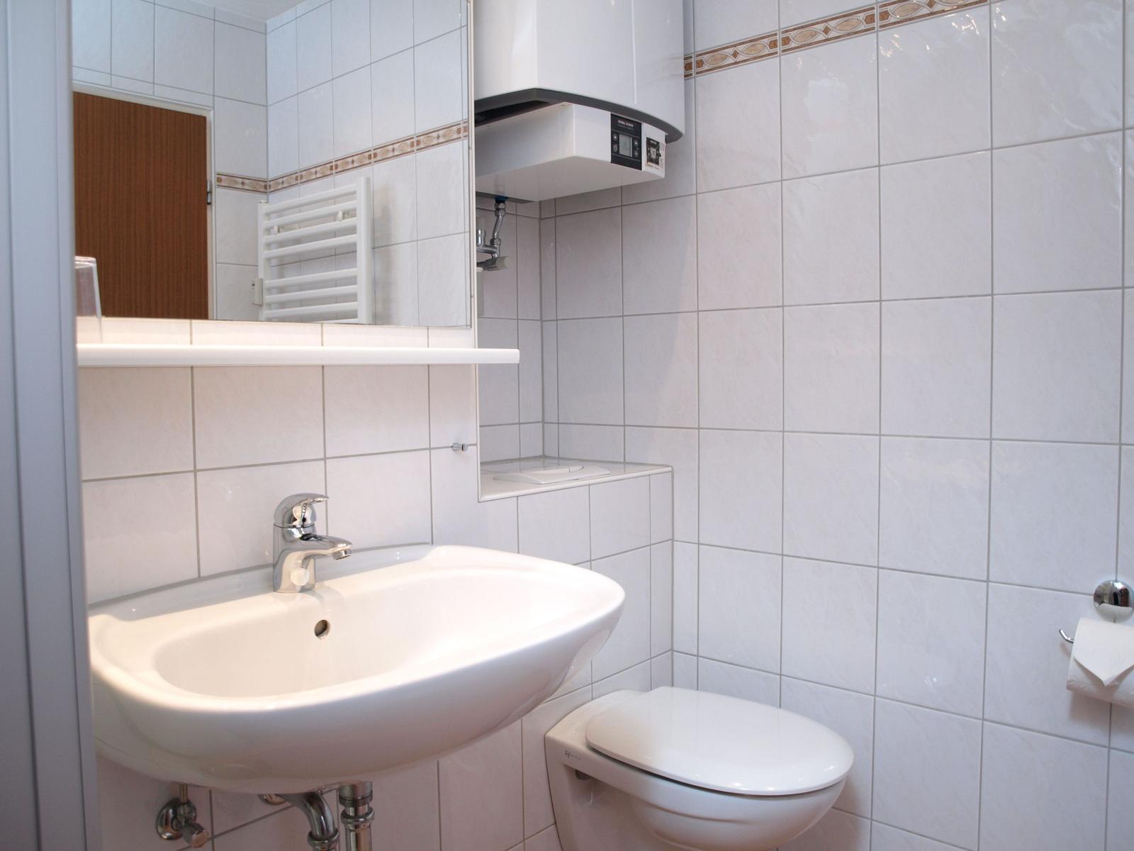Badezimmer mit Waschbecken, Toilette und Spiegel. Wandfliesen und Heizkörper sichtbar.