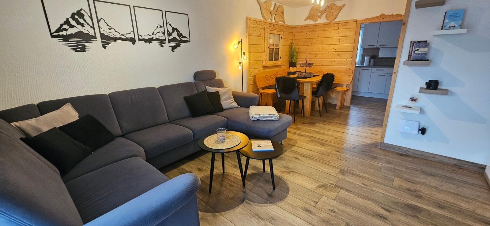 Wohnbereich mit Sofa, Esstisch und Küchenbereich. Holzdielenboden und Wanddekoration.