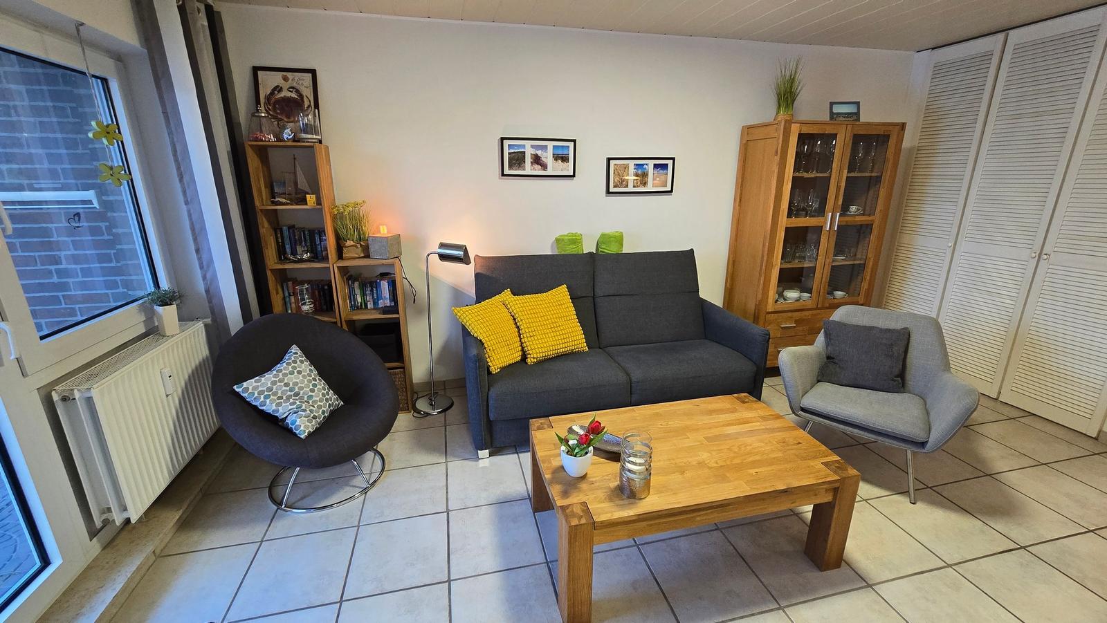 Wohnzimmer mit grauem Sofa, Holztisch und Bücherregal. Fenster und Heizung sichtbar.