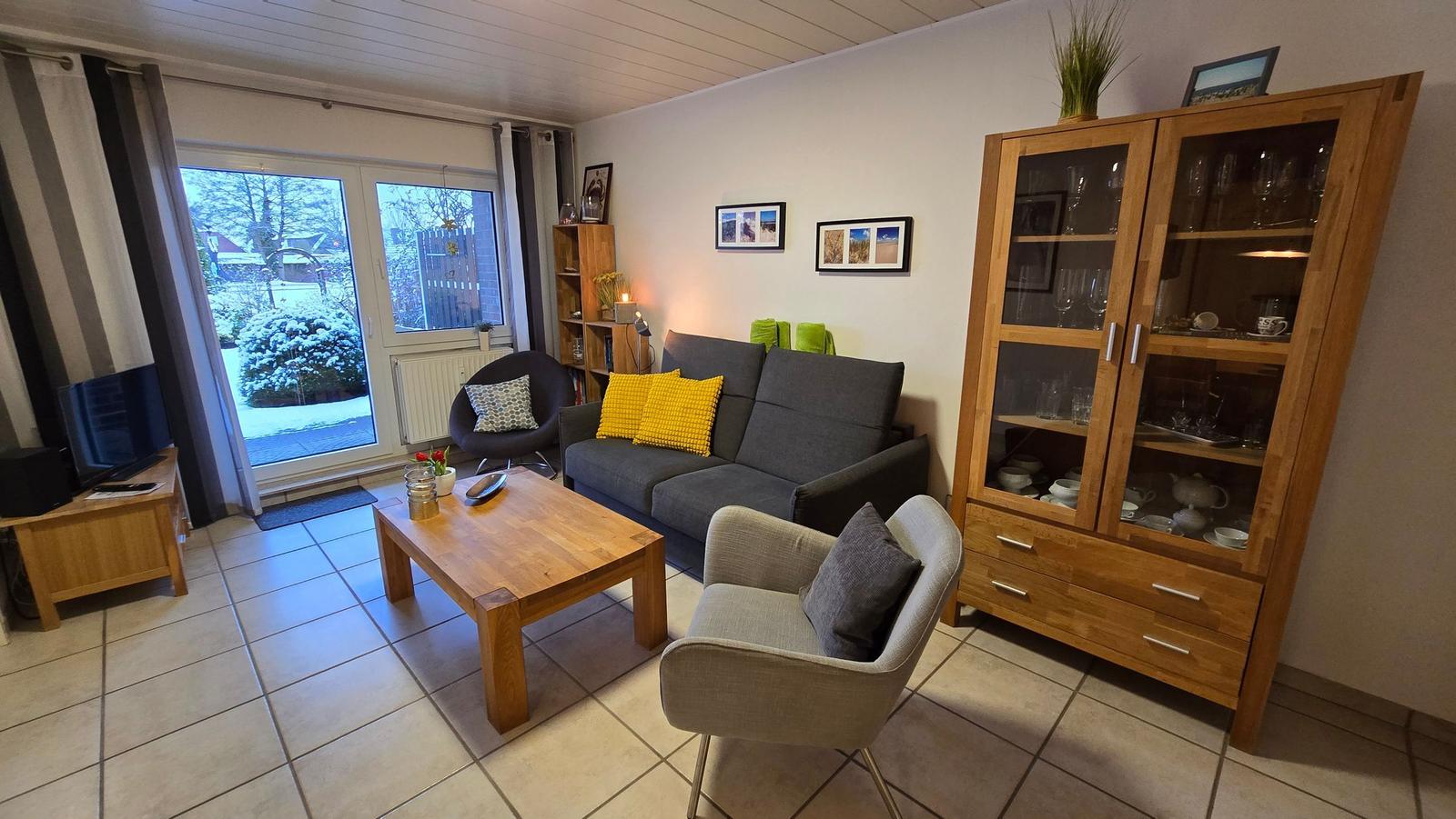 Wohnzimmer mit Sofa, Sessel und Glasvitrine. Durchgang zum Außenbereich mit Schnee.