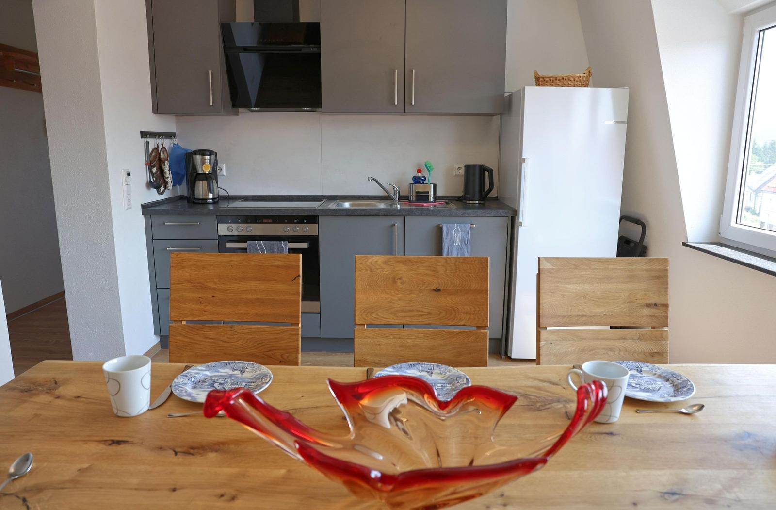 Küche mit Esstisch, drei Stühlen und rotem Glasbehälter. Fenster und Kühlschrank sind sichtbar.
