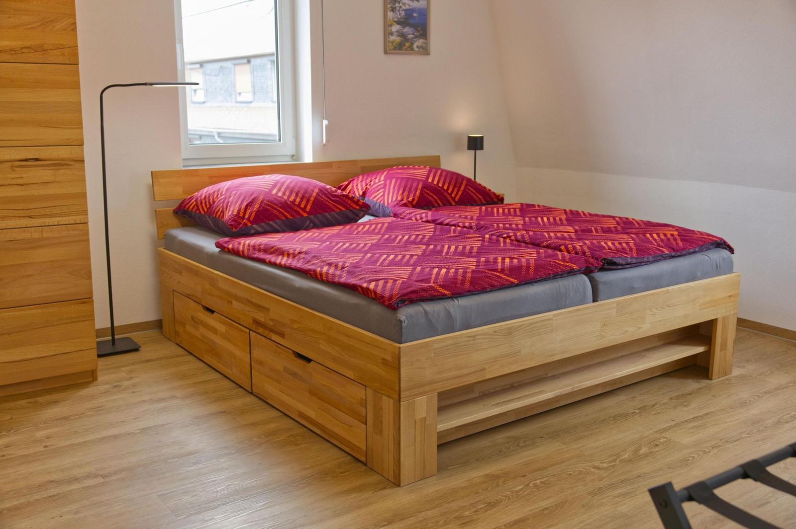 Doppelbett mit Holzrahmen und integrierten Schubladen, rote Bettwäsche, Fenster und Stehlampe.