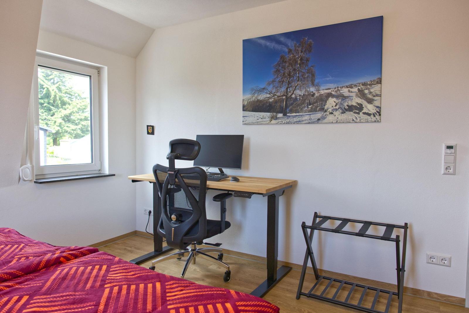Schlafzimmer mit Schreibtisch, Bett und Wandbild. Fenster mit Blick nach draußen.