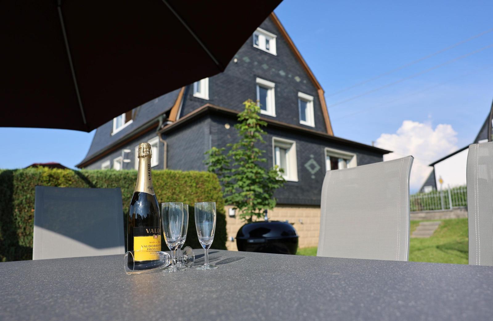 Außenbereich mit Tisch, Stühlen und einer Flasche Prosecco unter einem Sonnenschutz.