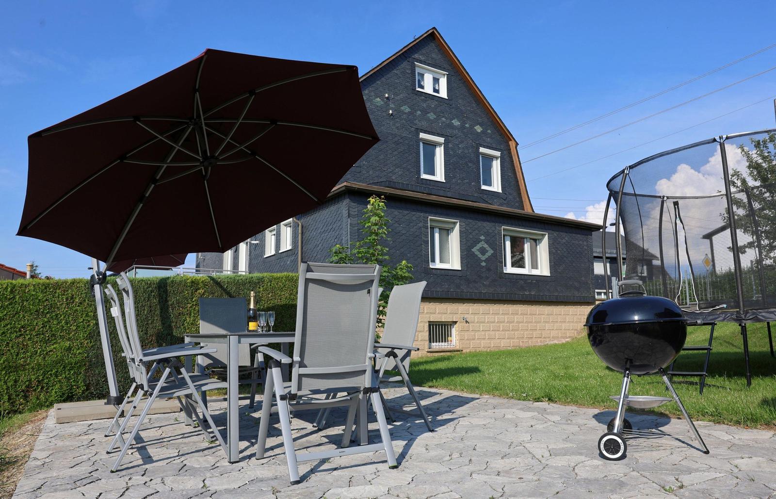 Terrasse mit Tisch, Stühlen, Sonnenschirm und Grill. Hintergrund: Haus mit Schieferdach.