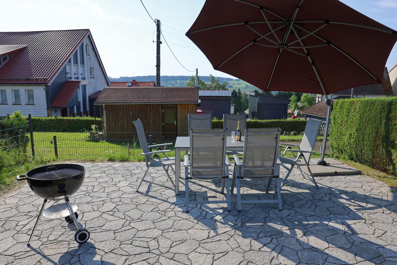 Terrasse mit Tisch, Stühlen, Sonnenschirm und Grill. Hintergrund: Haus und Landschaft.