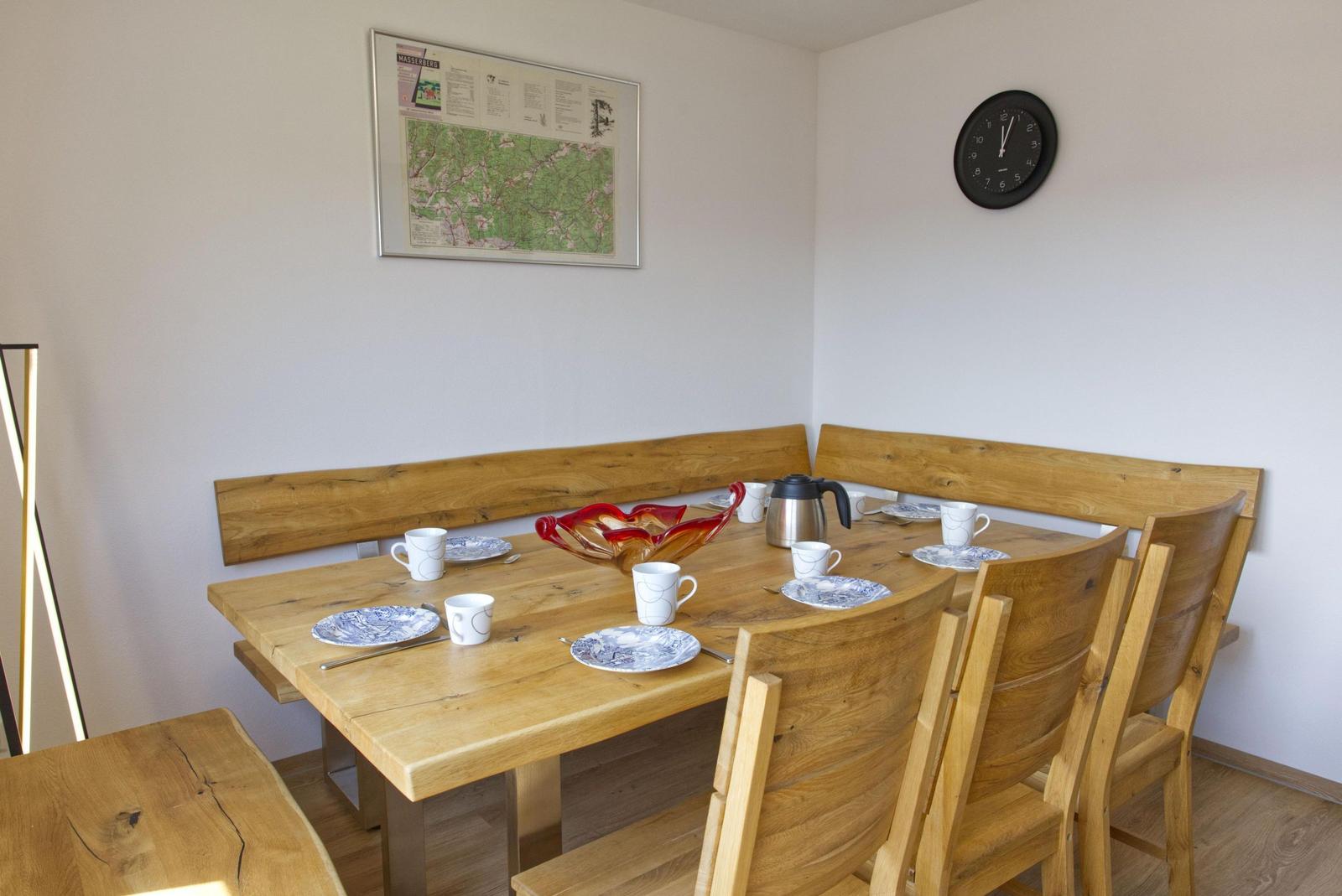 Esszimmer mit Holztisch, Bänken und Stühlen. Wanduhr und Wandkarte sind sichtbar.