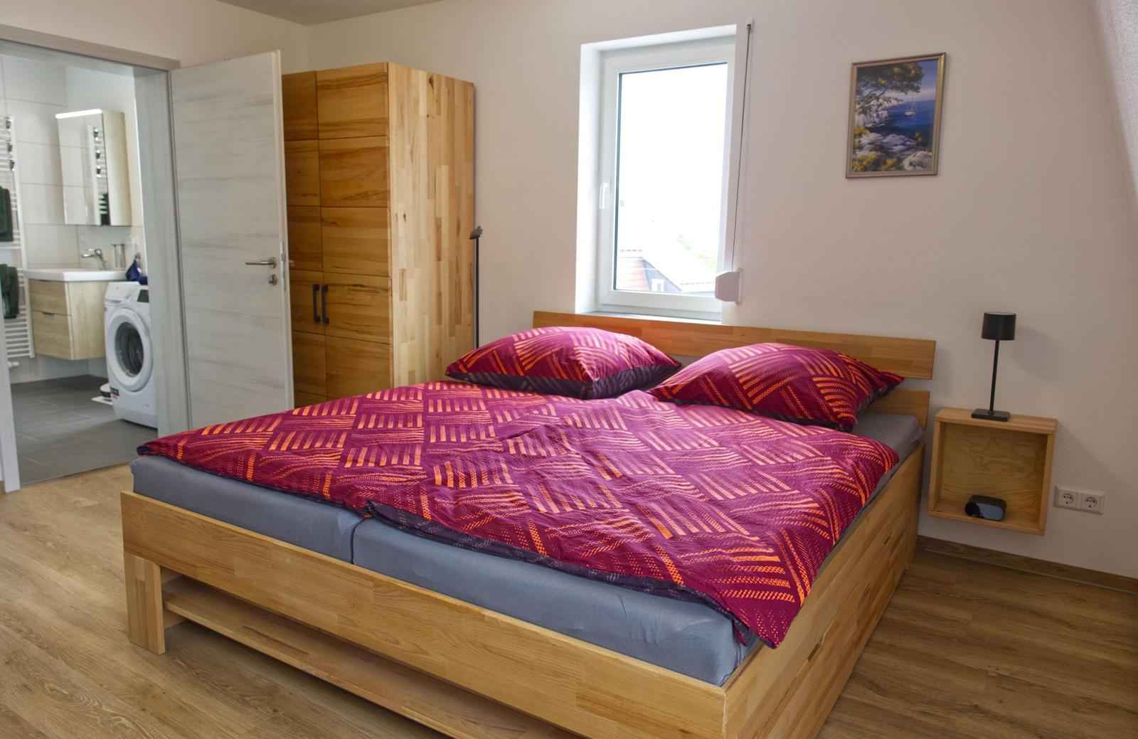 Schlafzimmer mit Holzbett, Kleiderschrank und Fenster. Nebenraum mit Waschmaschine und Spiegel.